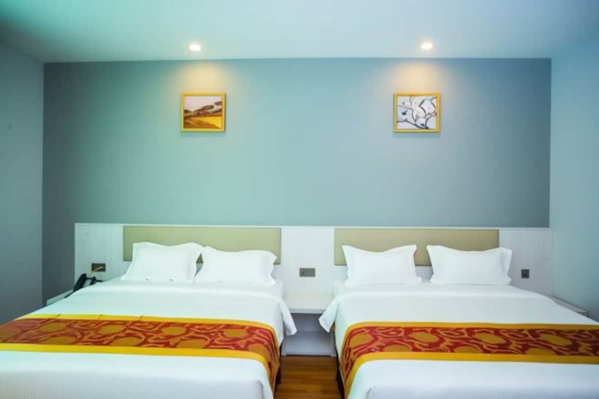 A hotel room with two beds, each with white bedding and a patterned bedspread. There are two wall-mounted artworks, one depicting a landscape and the other an abstract design.