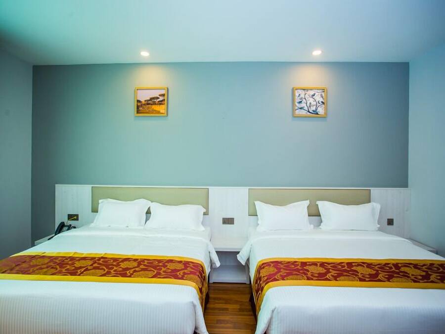 A hotel room with two beds, each with white linens and a red and gold patterned bedspread. The room has light blue walls, two framed artworks, and recessed ceiling lighting.