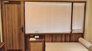 A room with a wooden door, a small bedside table with a lamp, a bed, and a large window with blinds. - Soleil House (Hue)