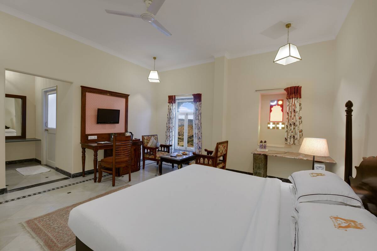 Une chambre d’hôtel comprenant un lit, un bureau, des chaises, une télévision, une fenêtre avec des rideaux, un ventilateur de plafond et une lampe.