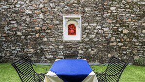 Un muro de piedra con una estatua enmarcada, una mesa cubierta con mantel azul y dos sillas de metal negro.