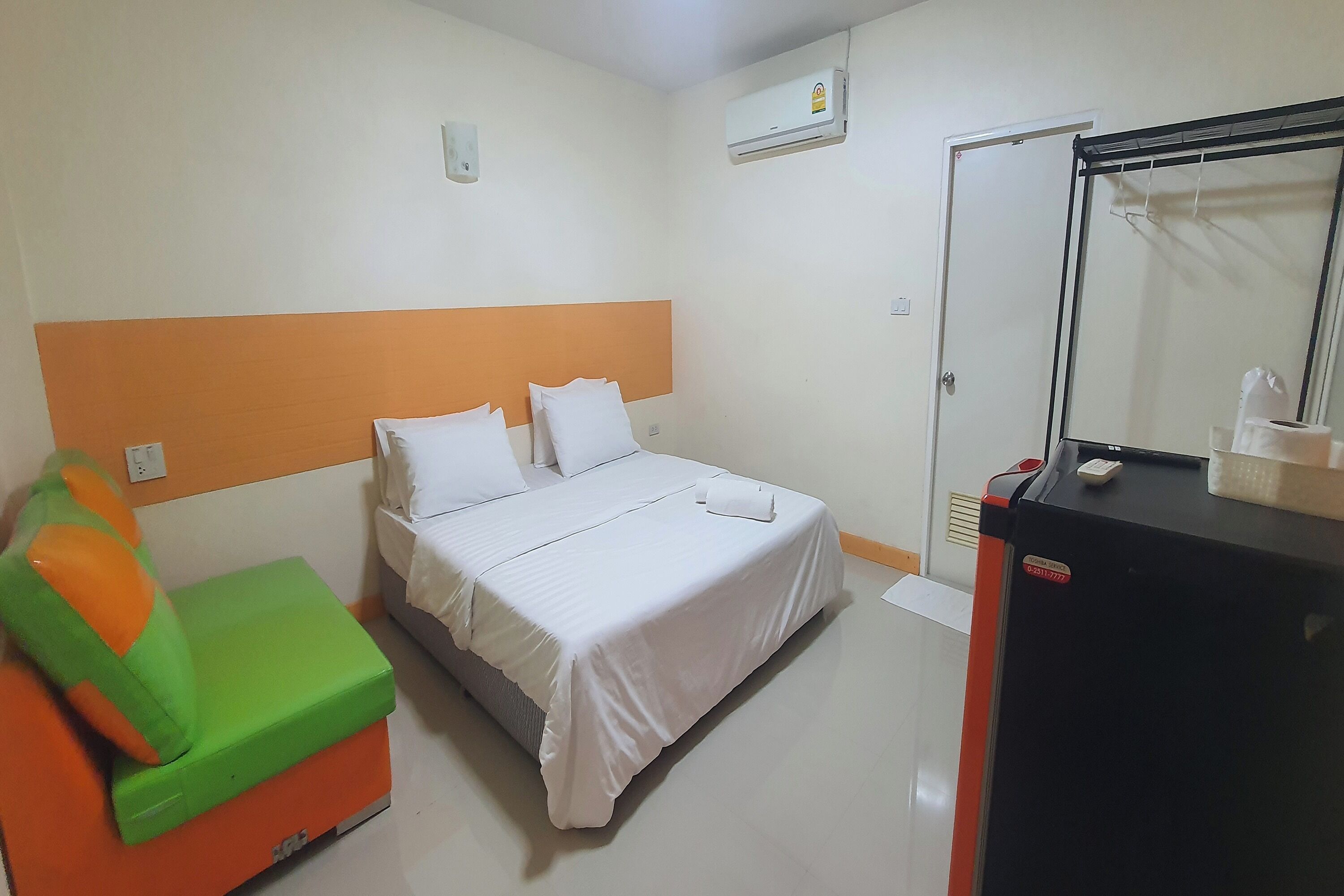A hotel room with a bed, a green and orange chair, a black minibar, and a mirror.