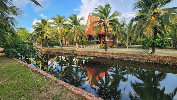 Uma casa tradicional com telhado de telhas, cercada por palmeiras e um corpo d'água calmo que reflete a construção.