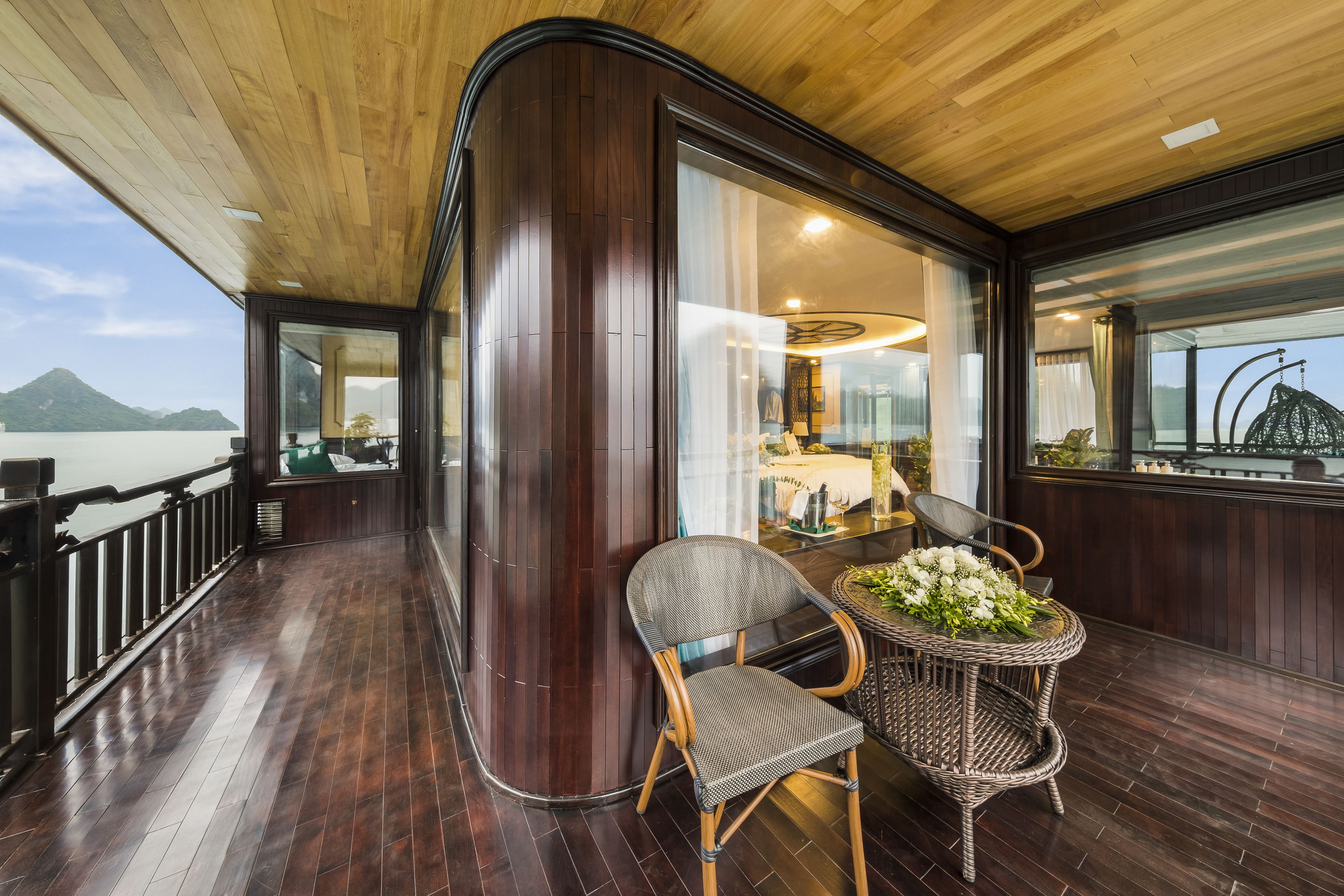 Una terraza de madera con vista a una montaña y al agua, un sillón de mimbre y una mesa con flores.