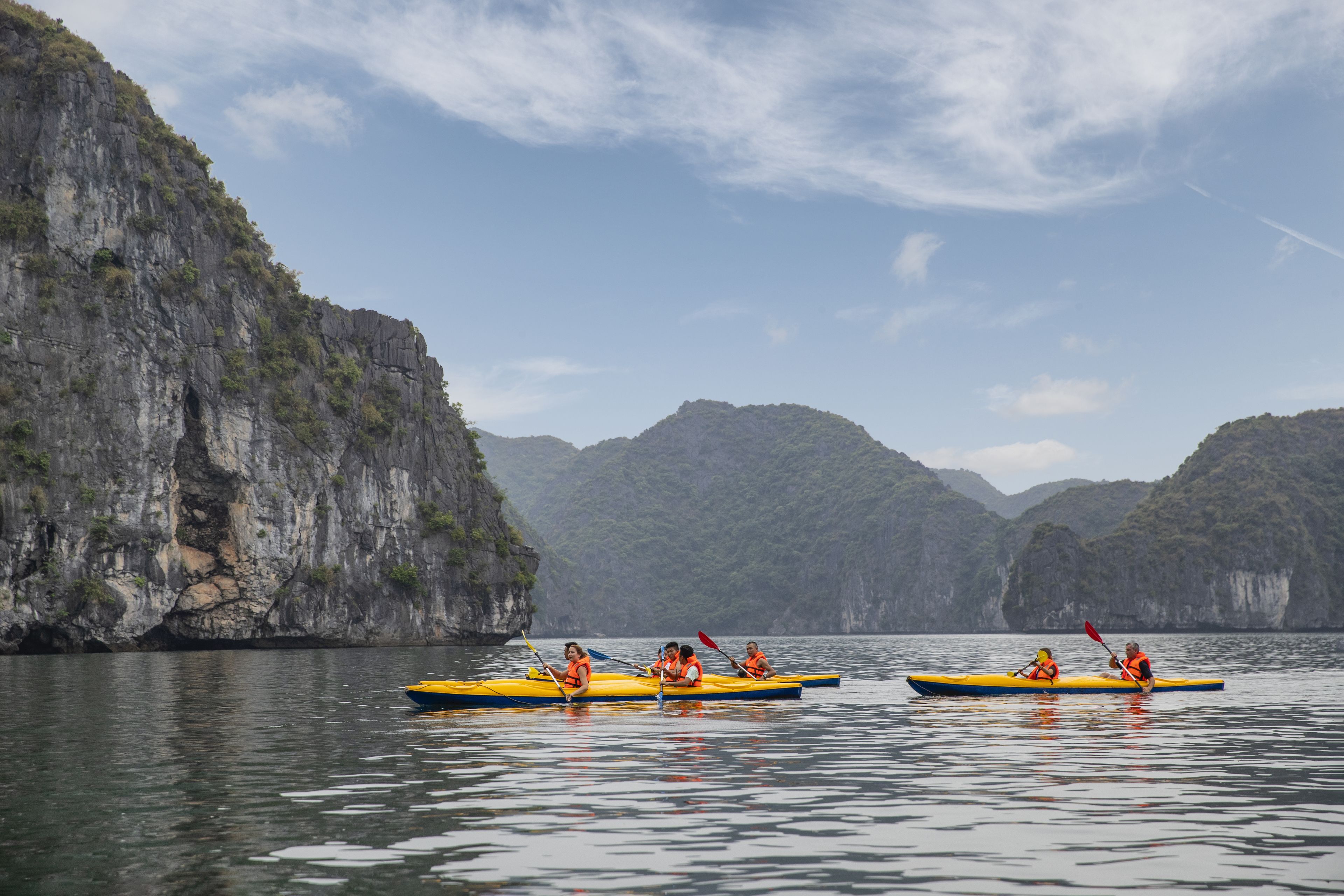 A group of people kayaking on a calm body of water with rocky cliffs in the background.