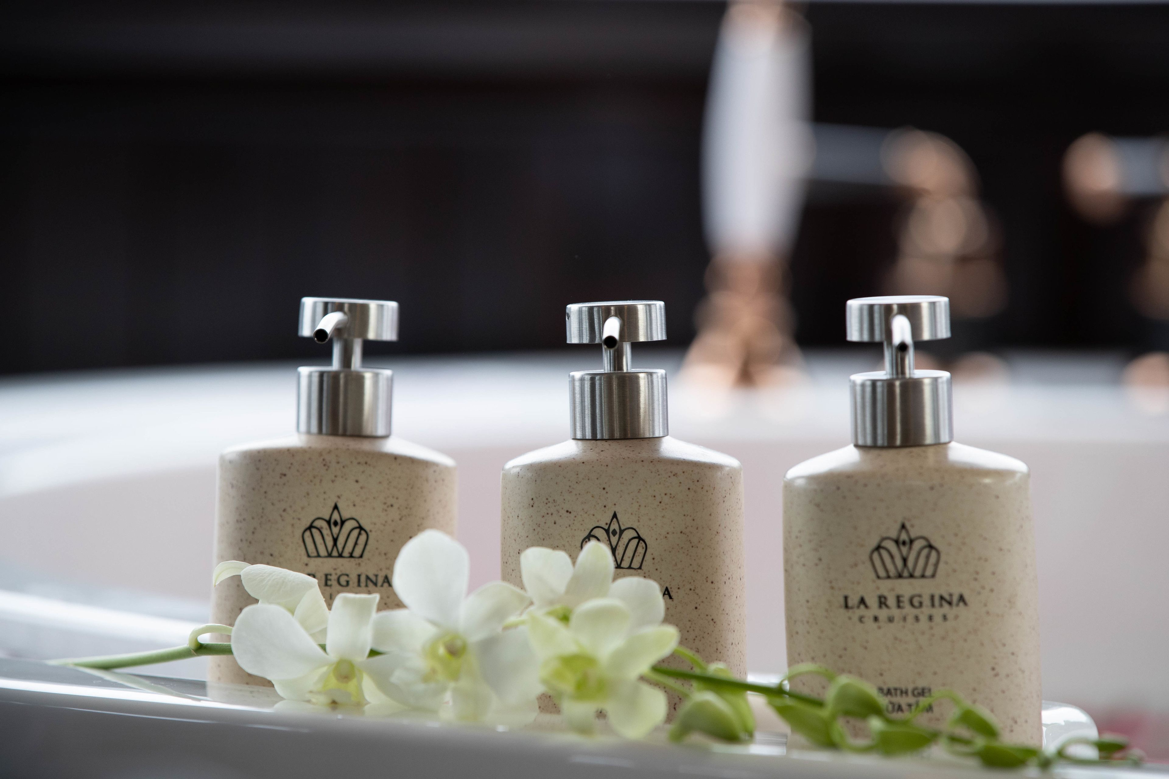 Three La Regina Cruises bath gel bottles on a white surface with white orchids.