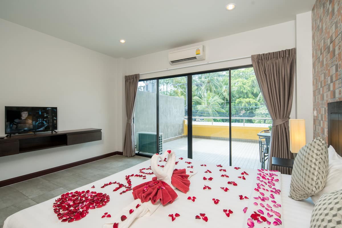 Une chambre d’hôtel avec un lit parsemé de pétales de rose et décoré d’un cœur, un téléviseur posé sur un meuble, et un balcon offrant une vue sur la verdure.