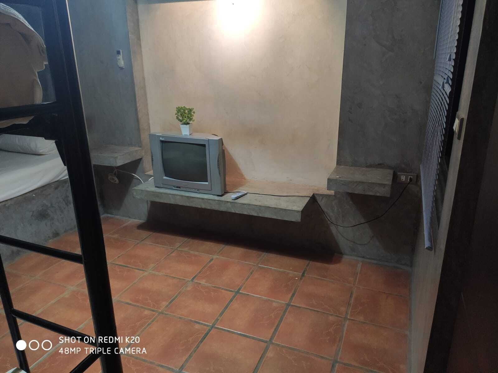 Una habitación con piso de baldosas, un televisor pequeño en una repisa y una cama con colchón blanco.