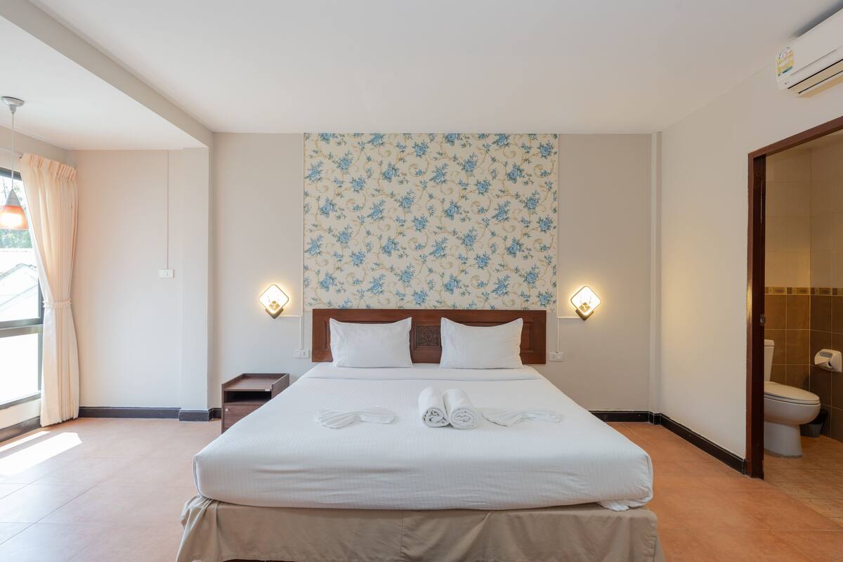 Une chambre d’hôtel avec un grand lit, deux lampes de chevet, un papier peint à motifs floraux et une salle de bain avec des toilettes et un lavabo.