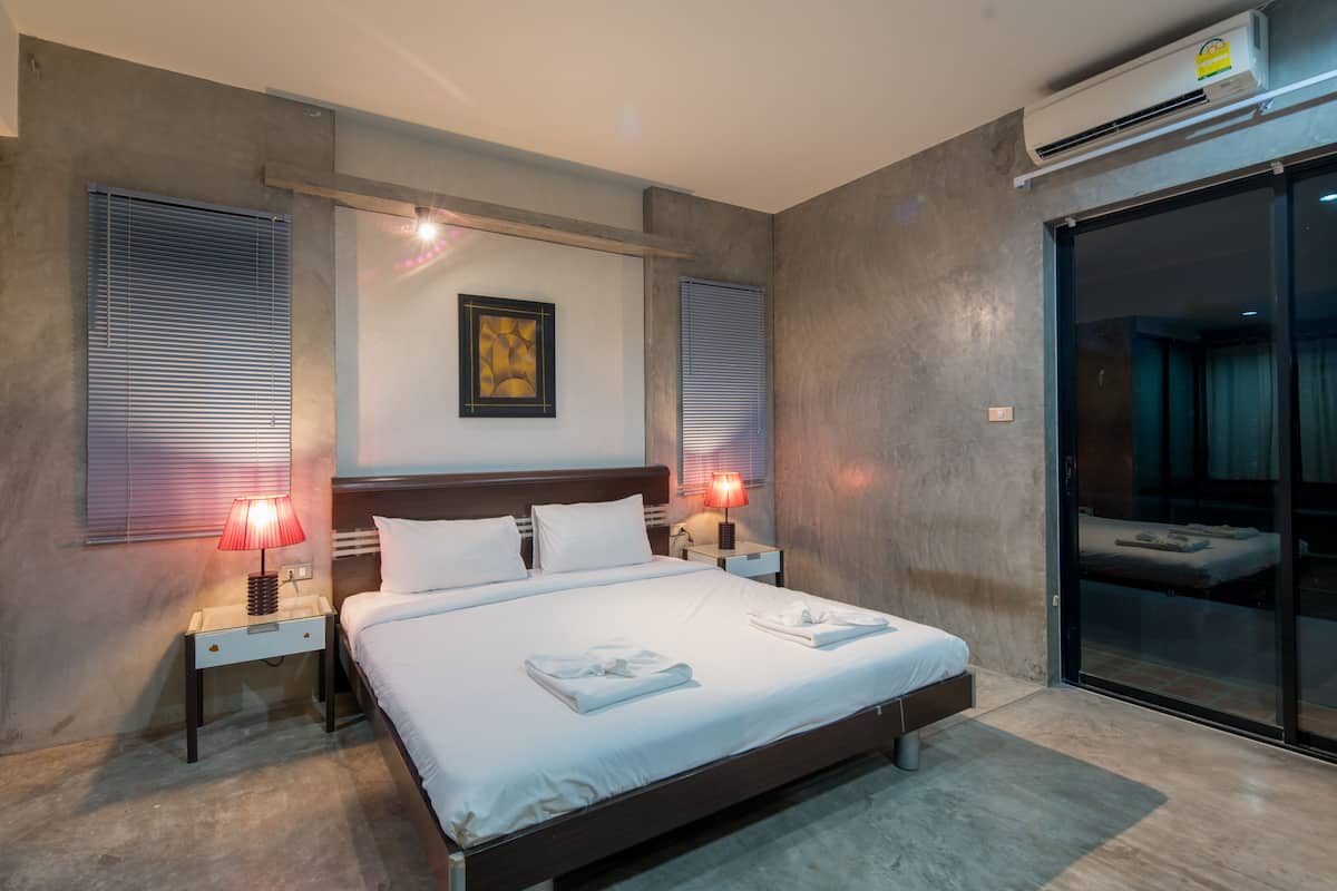 Une chambre d’hôtel moderne dotée d’un grand lit, de deux lampes de chevet, d’un climatiseur mural et d’une porte-fenêtre coulissante donnant sur le balcon.