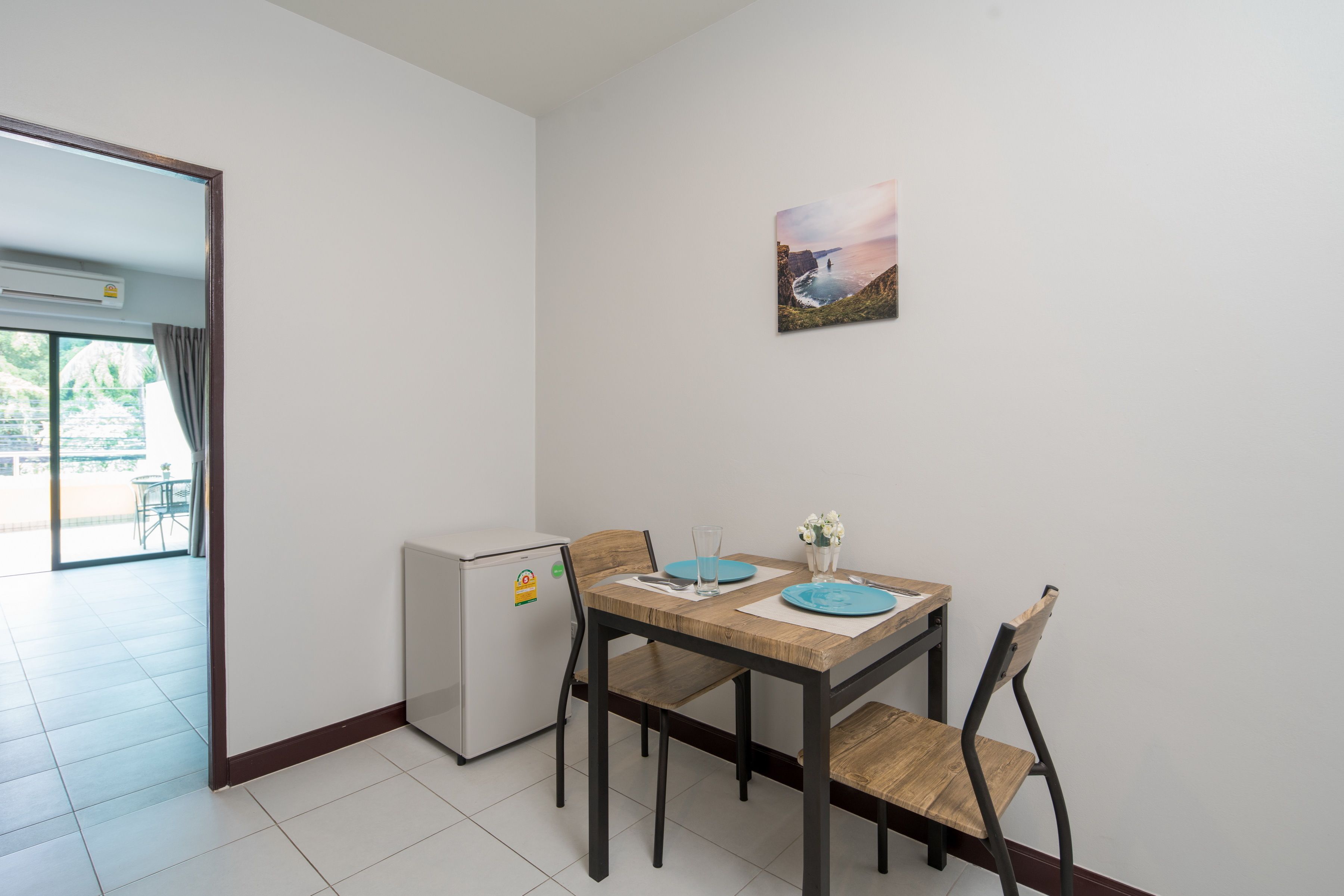 A dining area with a wooden table, two chairs, a refrigerator, and a wall-mounted picture.