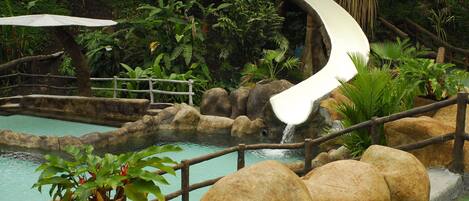 A water slide in a tropical setting with a pool, rocks, and lush greenery.