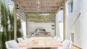 Une terrasse extérieure avec une table en bois et des chaises blanches, un barbecue et une pergola agrémentée de guirlandes lumineuses.