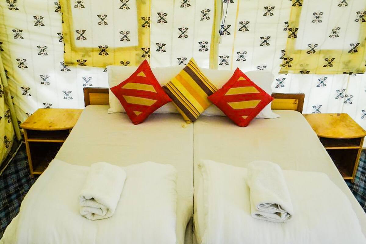 Un lit double avec deux oreillers, une petite table et une chaise dans une pièce dont les fenêtres sont ornées de rideaux à motifs.