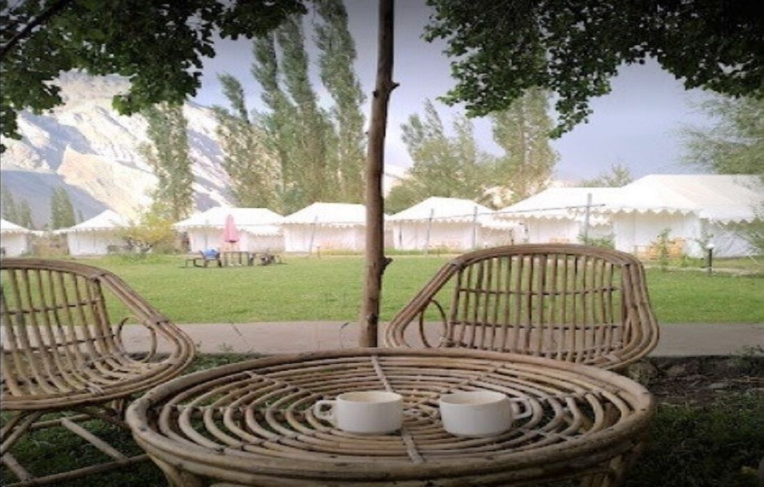A rustic outdoor seating area with wicker chairs and a table, overlooking a grassy field with white tents in the background.