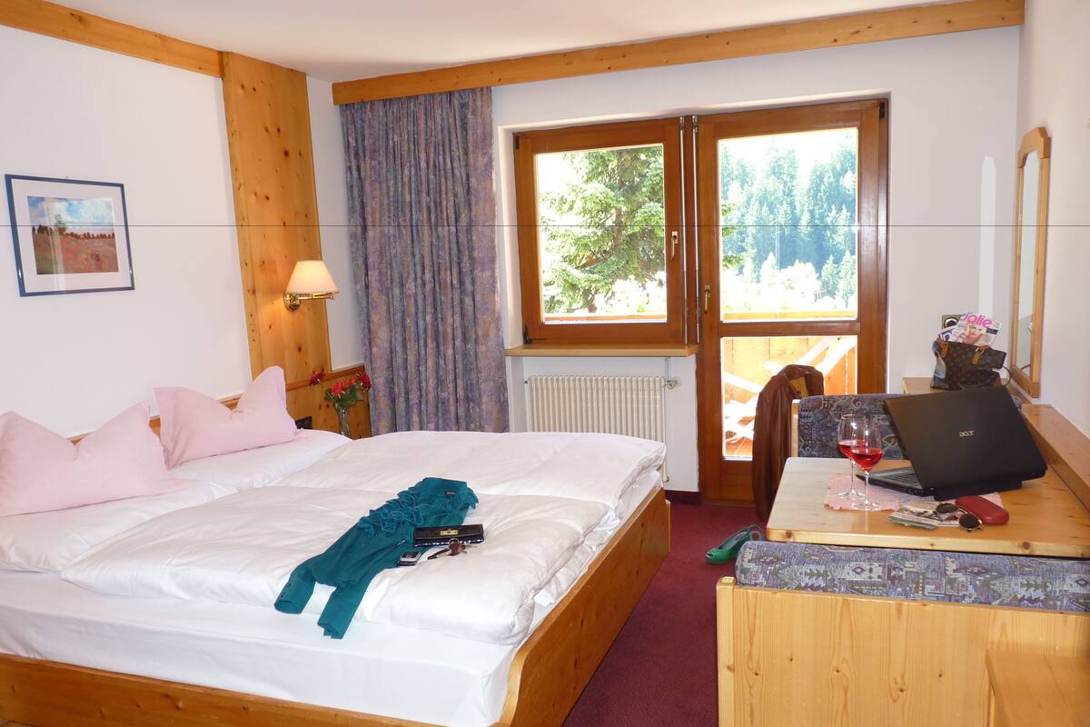 A wooden-framed room with a bed, a desk with a laptop, and a window with a view of trees.
