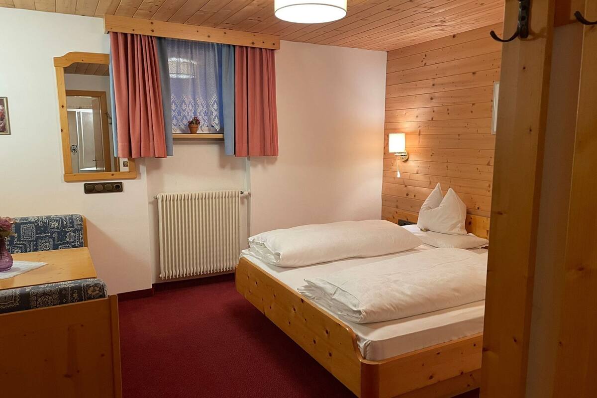 A wooden-paneled room with a bed, a radiator, a mirror, a window with curtains, and a small table with a vase.