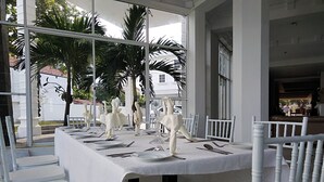 A dining area with white table settings and chairs, large windows, and a view of palm trees and a building outside. - Cinnamon Villa Negombo (Negombo)