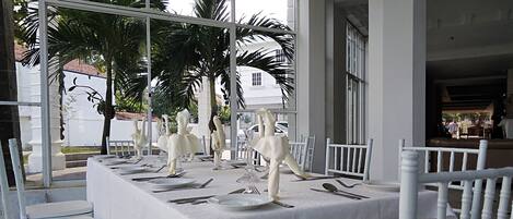 Un comedor con vajilla blanca, sillas, ventanas amplias y vistas a palmeras y un edificio.