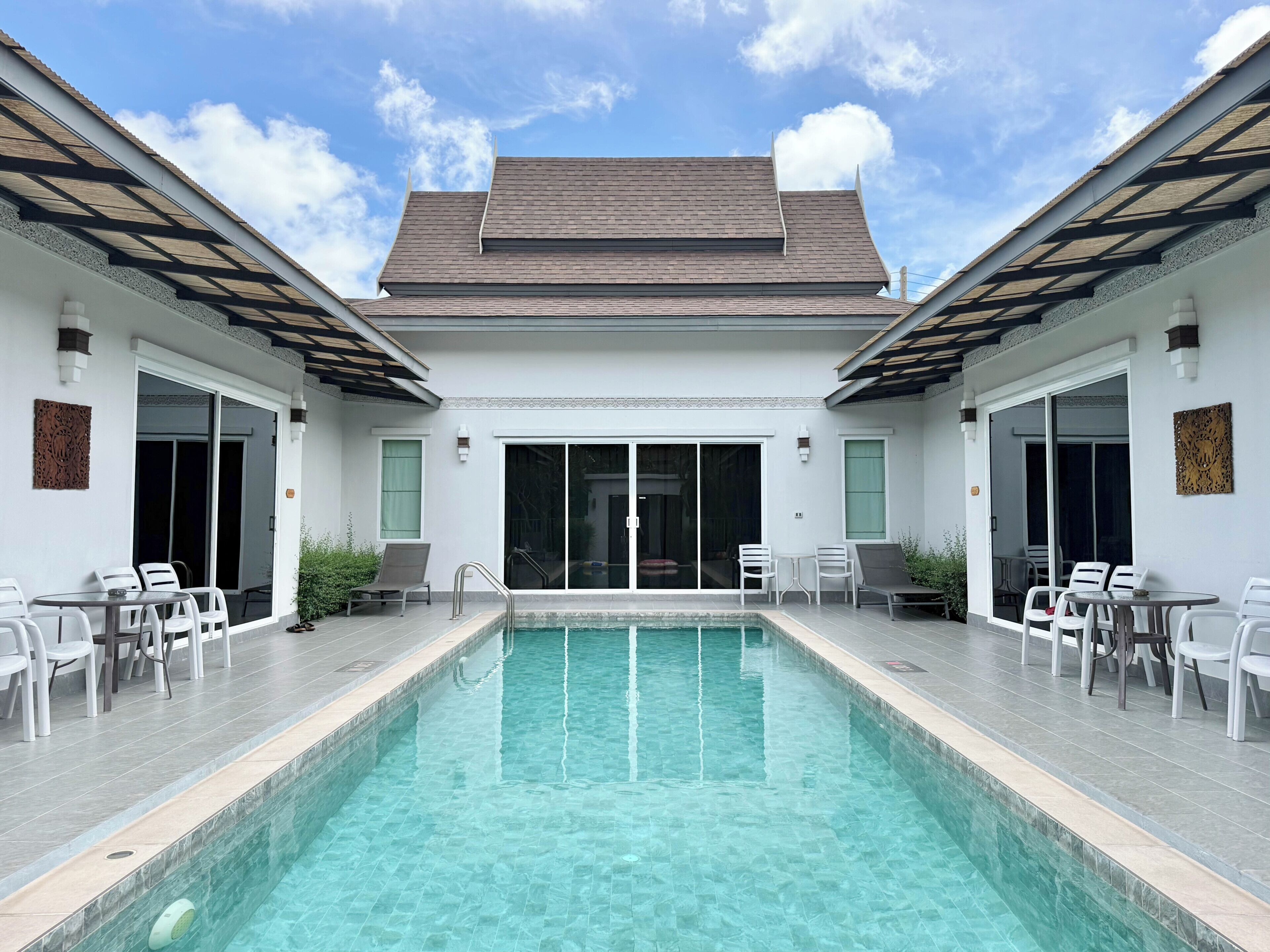 Un área de piscina con piscina transparente, sillas blancas y un porche cubierto.