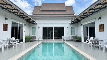 Un área de piscina con piscina transparente, sillas blancas y un porche cubierto.