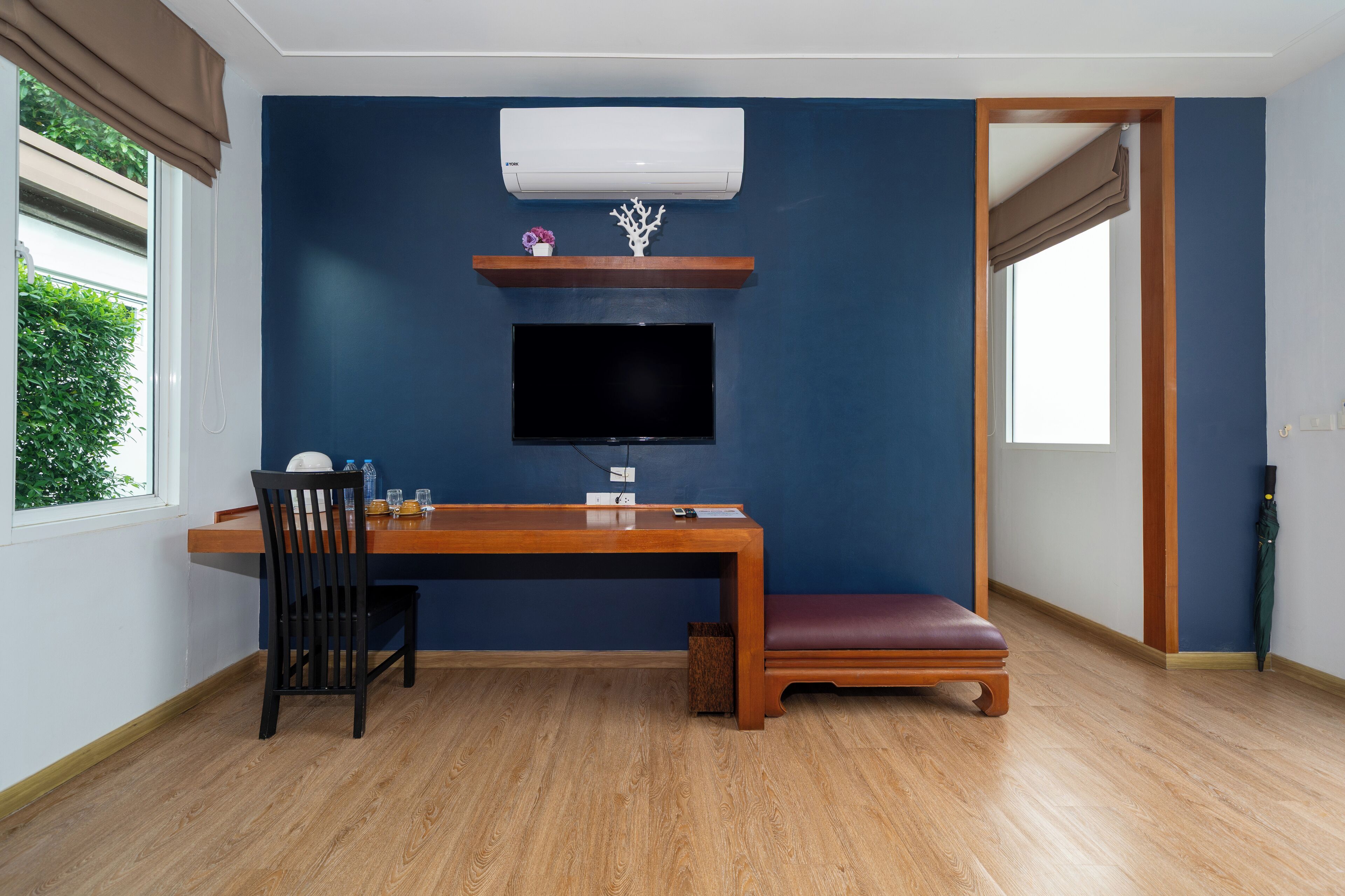 A room with a wooden desk, a chair, a television mounted on the wall, a bench, and a window with curtains.