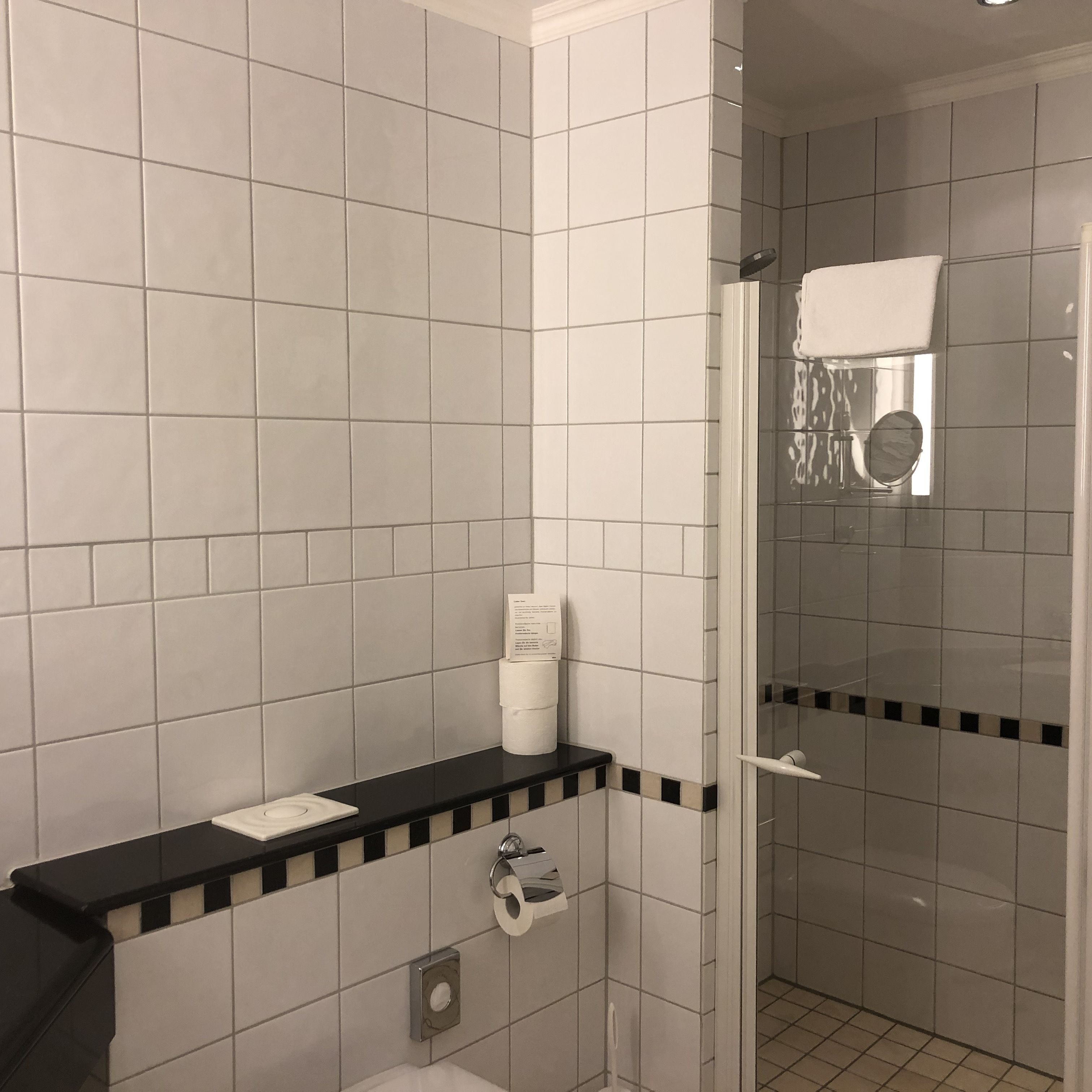 A bathroom with white tiles, a black countertop, a toilet, a paper towel dispenser, and a glass shower enclosure.