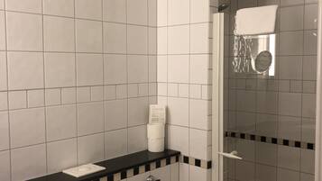 A bathroom with white tiles, a black countertop, a toilet, a paper towel dispenser, and a glass shower enclosure.