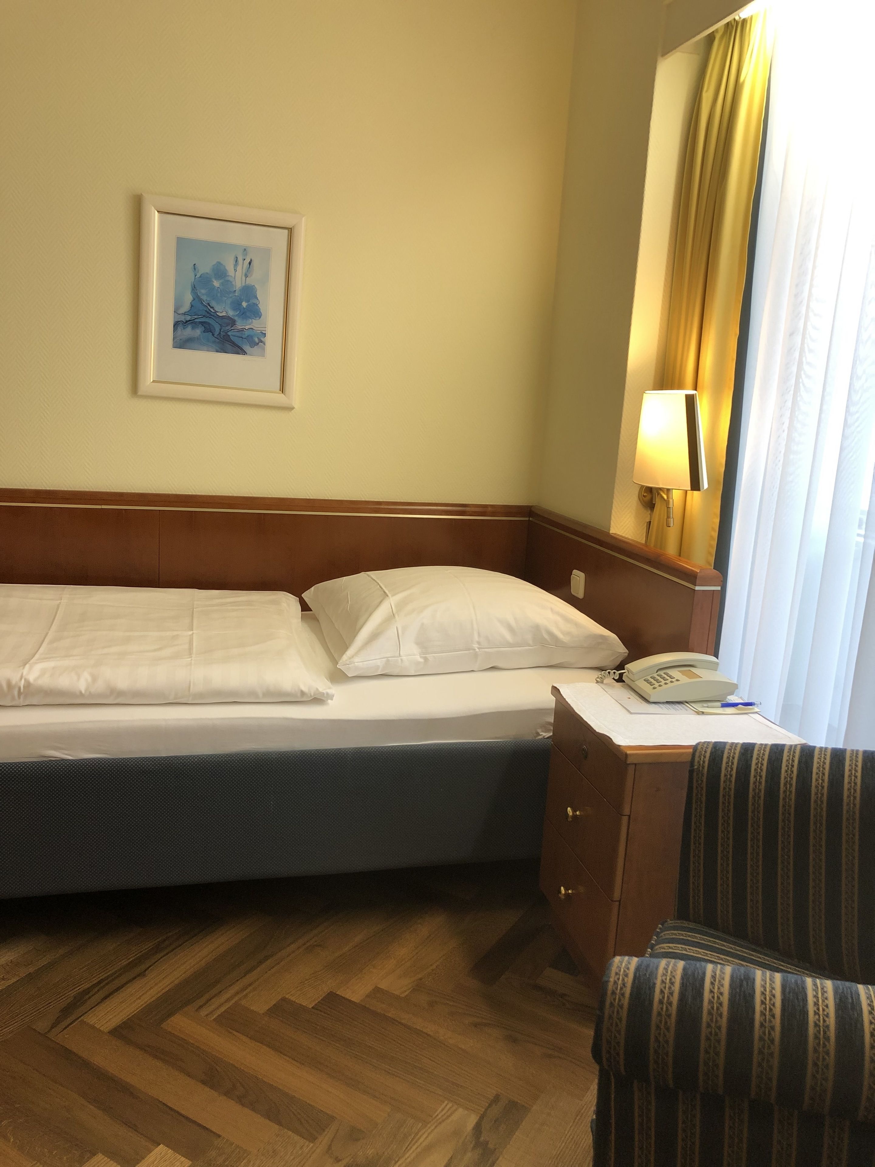 Ett hotellrum med en säng, ett nattduksbord med en lampa, en stol och en inramad tavla på väggen.