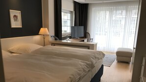 Habitación de hotel con una cama grande, un escritorio con computadora, una silla y una ventana con cortinas.