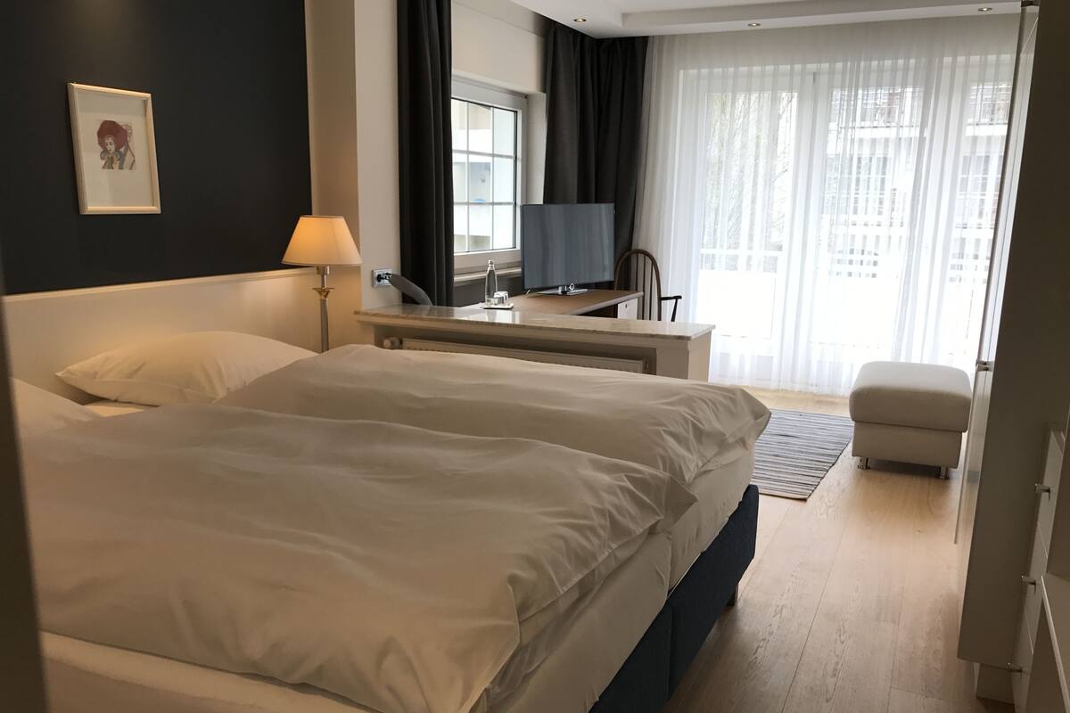 Quarto de hotel com uma cama grande, uma escrivaninha com computador, uma cadeira e uma janela com cortinas.