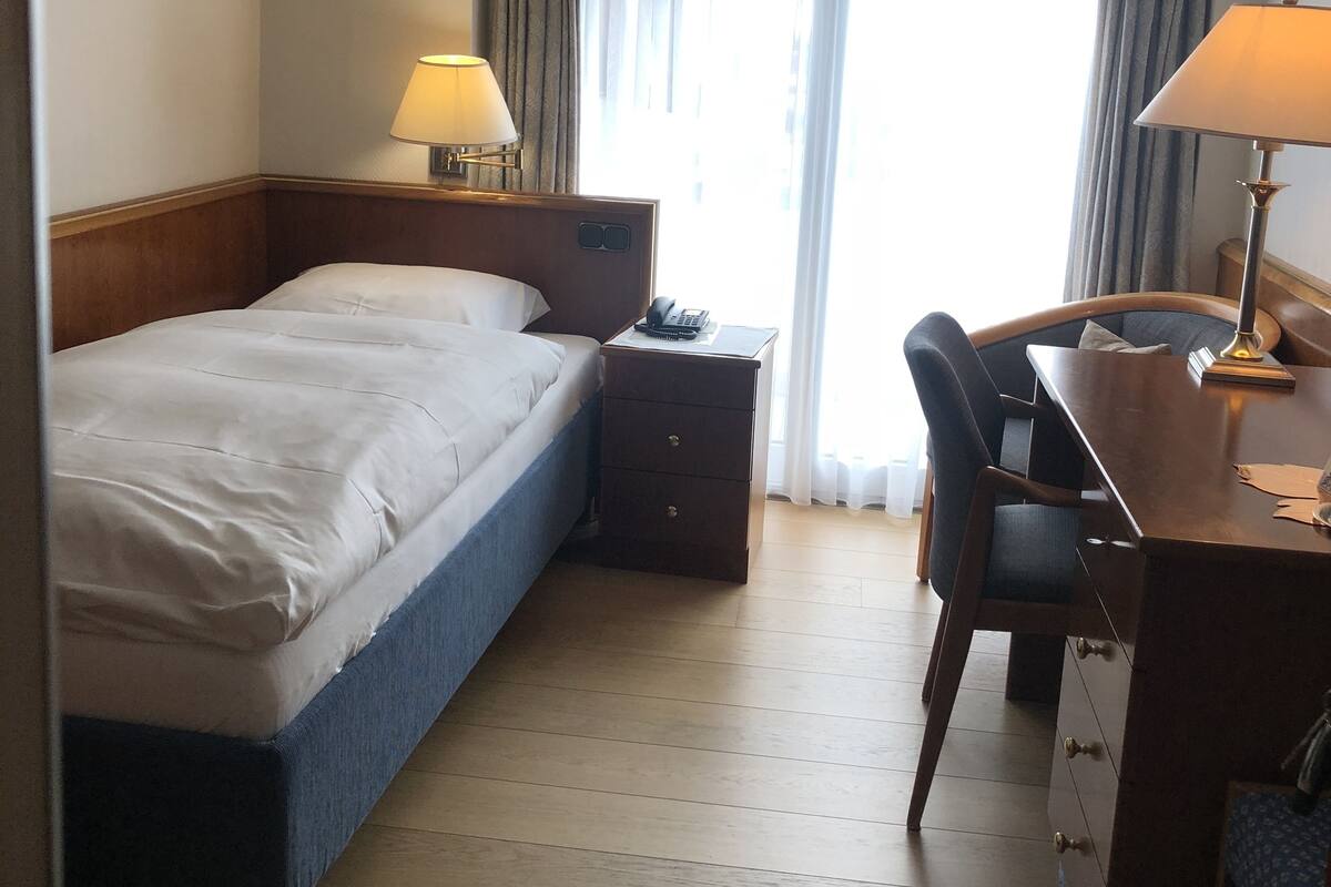 Quarto de hotel com cama, mesa de cabeceira, luminária, cadeira, escrivaninha e vista pela janela.