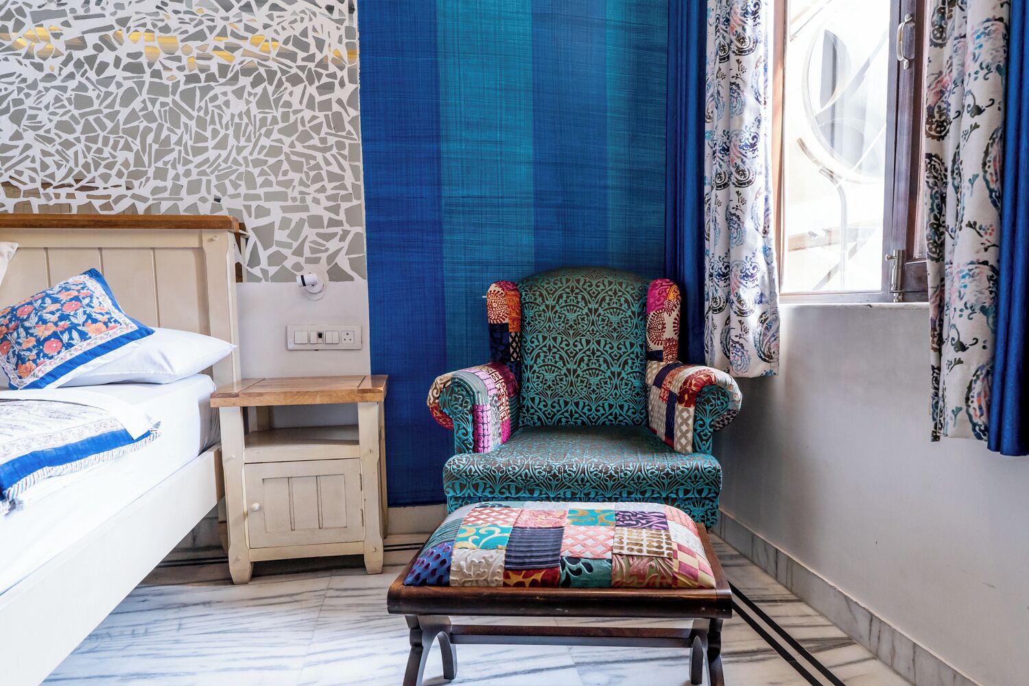 Un dormitorio con cama, mesita de noche, sillón con colcha y ventana con cortinas.