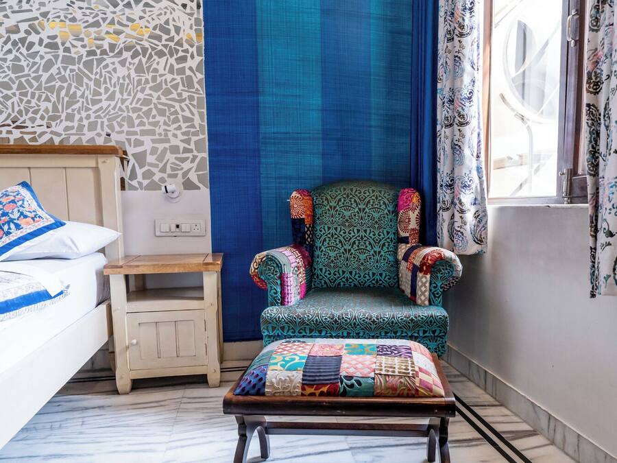 Un dormitorio con cama, mesita de noche, sillón con colcha y ventana con cortinas.