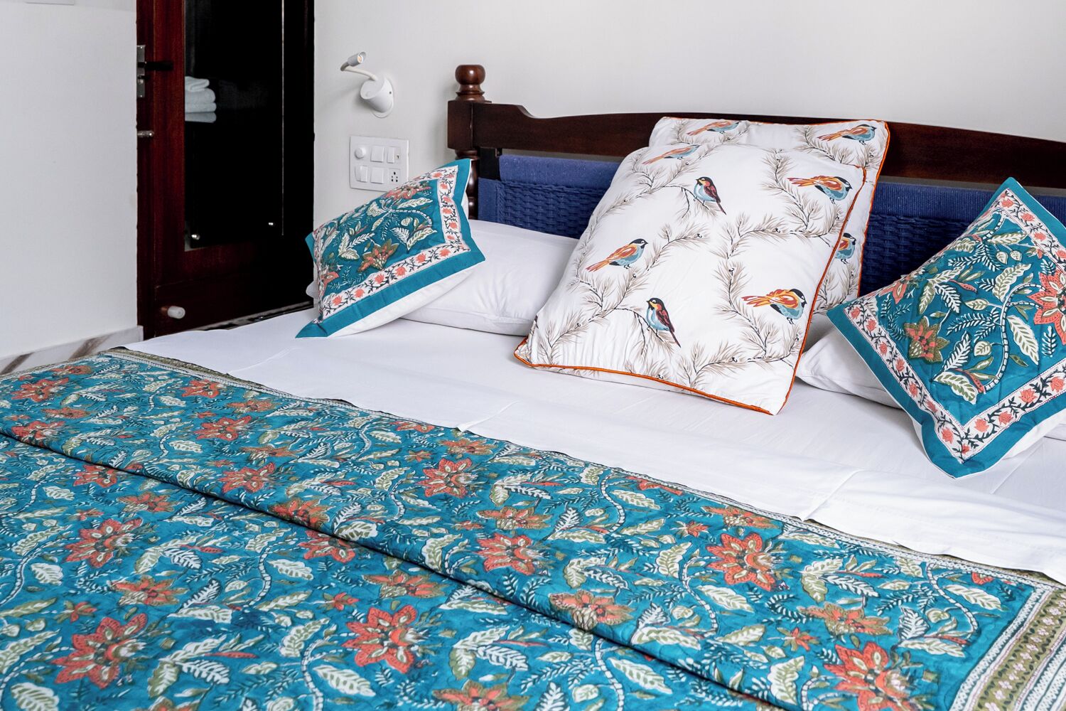 Un lit bien fait, avec une couvre-lit à motifs floraux et des coussins assortis, une tête de lit en bois et un luminaire fixé au mur.