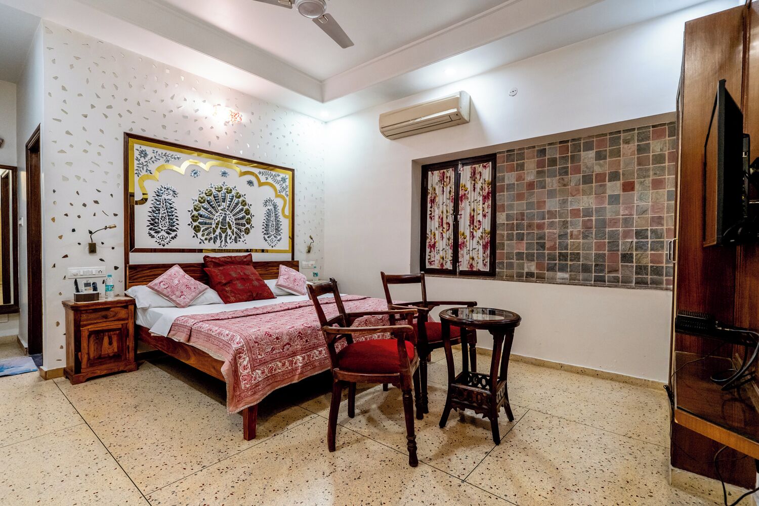 A bedroom with a bed, two chairs, a small table, a nightstand, a ceiling fan, and a wall-mounted air conditioner.