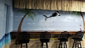Restaurante com um mural de um tubarão e palmeiras, cadeiras pretas e um telhado de palha.