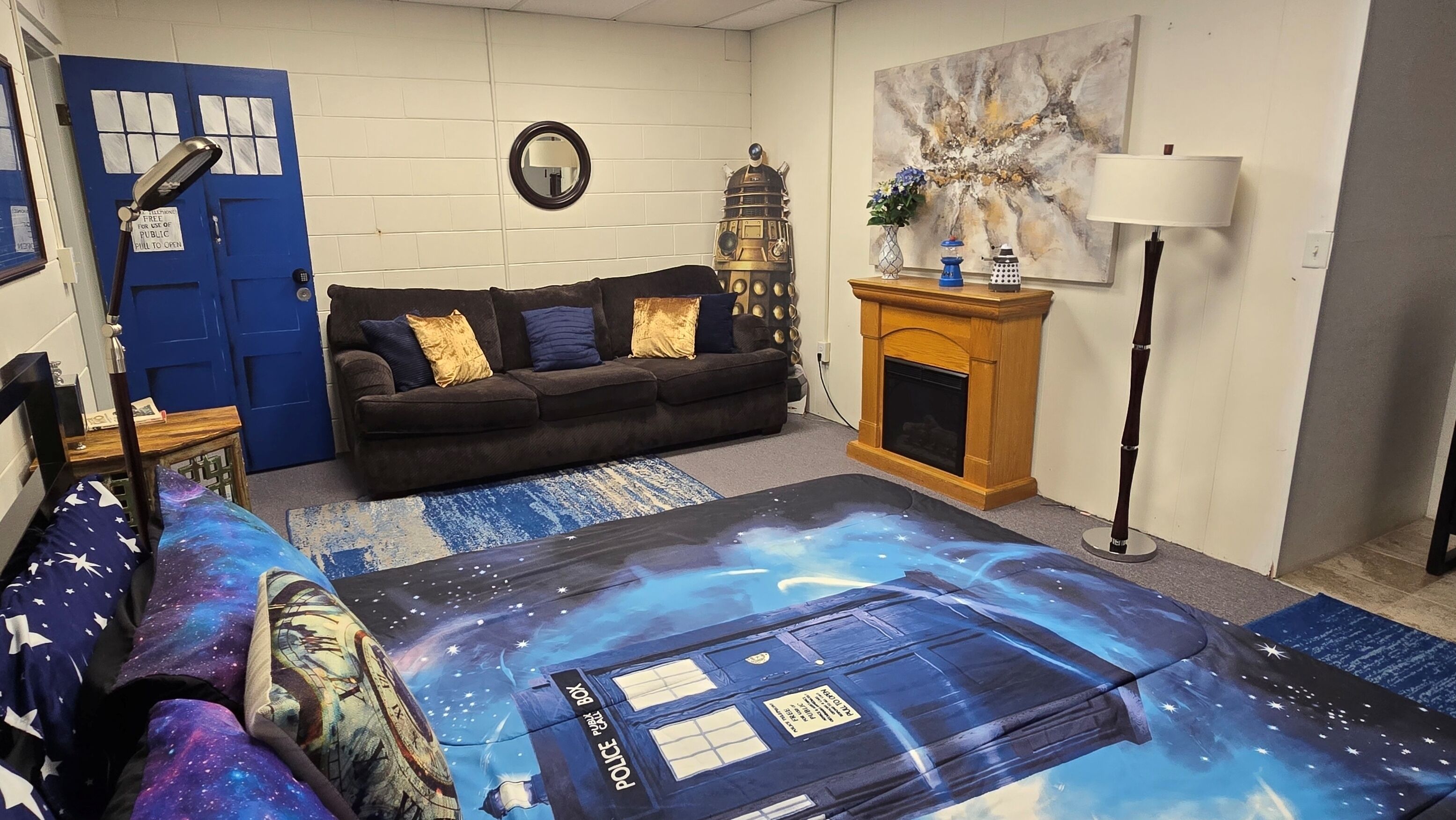 A room with a blue door, a sofa with patterned cushions, a wall-mounted mirror, a small table with a lamp, a fireplace, and a floor with a space-themed design.