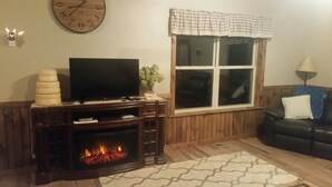 A living room with a fireplace, a television on a wooden stand, a window with a checkered valance, and a black leather sofa.
