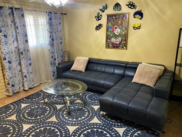 Un salon avec un canapé en cuir noir, une table basse en verre, un tapis à motifs, des rideaux à fleurs et un mur orné de masques décoratifs.