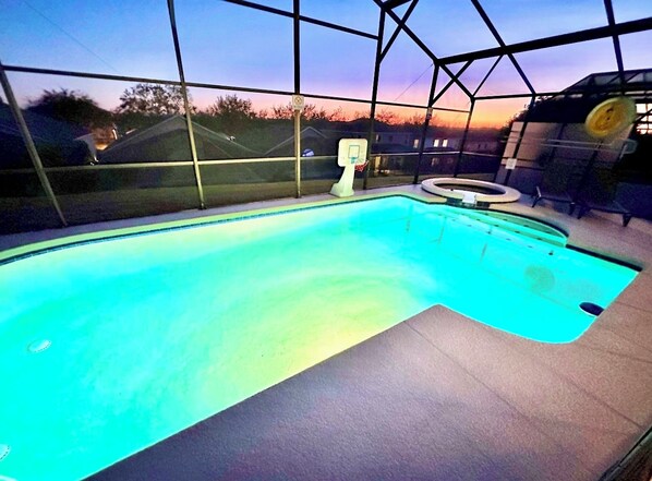 An indoor swimming pool with a basketball hoop and a view of the sunset through the glass enclosure.