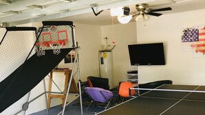 A room with a ping pong table, a basketball hoop, a chair, a TV, and a ceiling fan. - Perfect Vacation Getaway-Modern Gated Community, Spacious, Pool, Game Room, WiFi (Davenport)