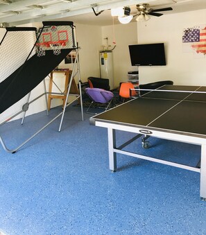 A room with a ping pong table, a basketball hoop, a chair, a TV, and a ceiling fan.