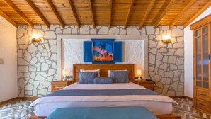 A bedroom with a stone wall, a wooden ceiling, a bed with blue bedding, and a nightstand with a lamp. - ADELLA GARDEN MARİNA HOTEL (Kusadasi)