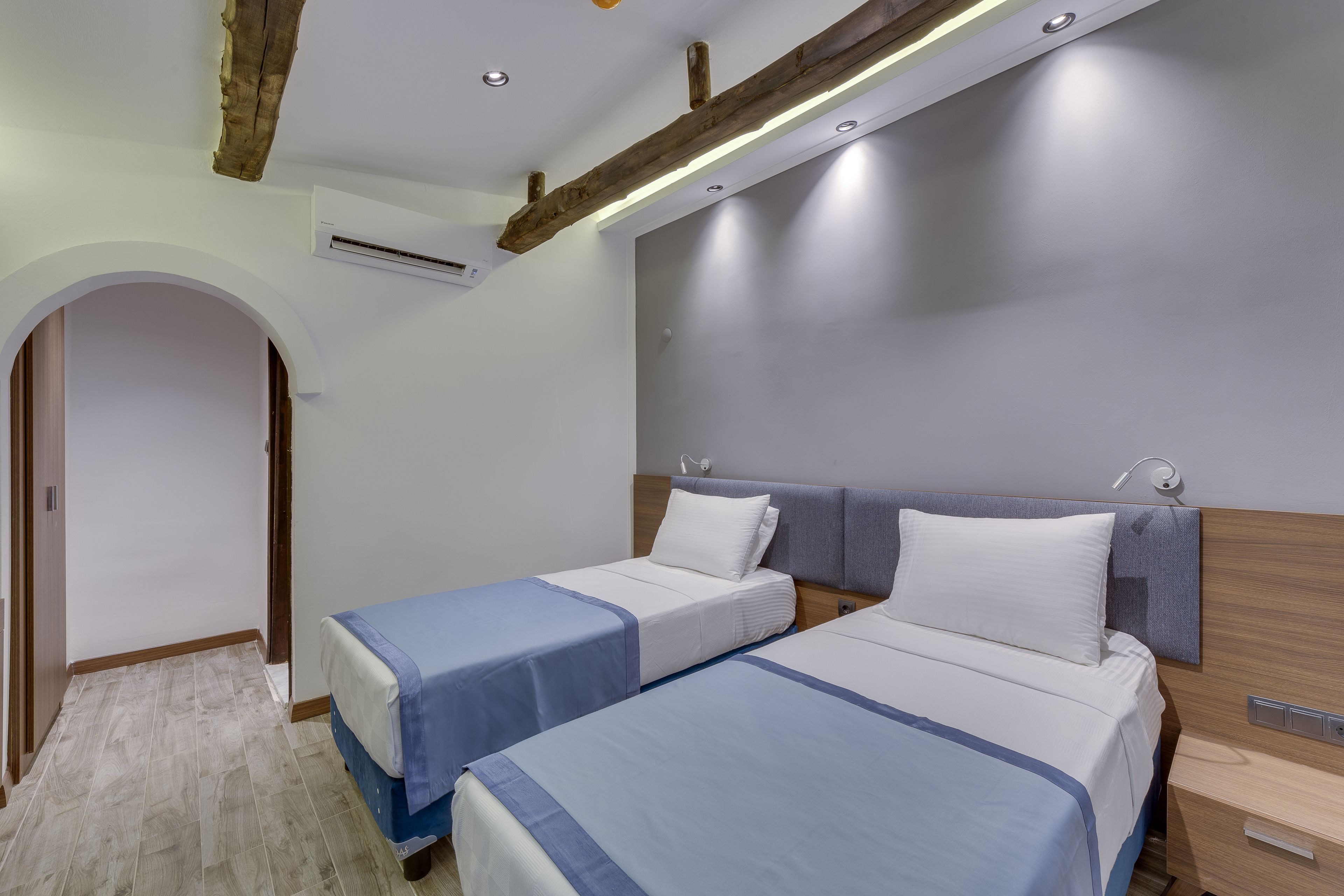 A hotel room with two beds, a wooden ceiling beam, an air conditioning unit, and a small bedside table.