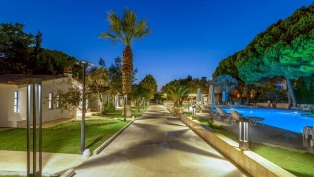 A pathway leading to a pool area with palm trees and a well-lit building on the left. - ADELLA GARDEN MARİNA HOTEL (Kusadasi)