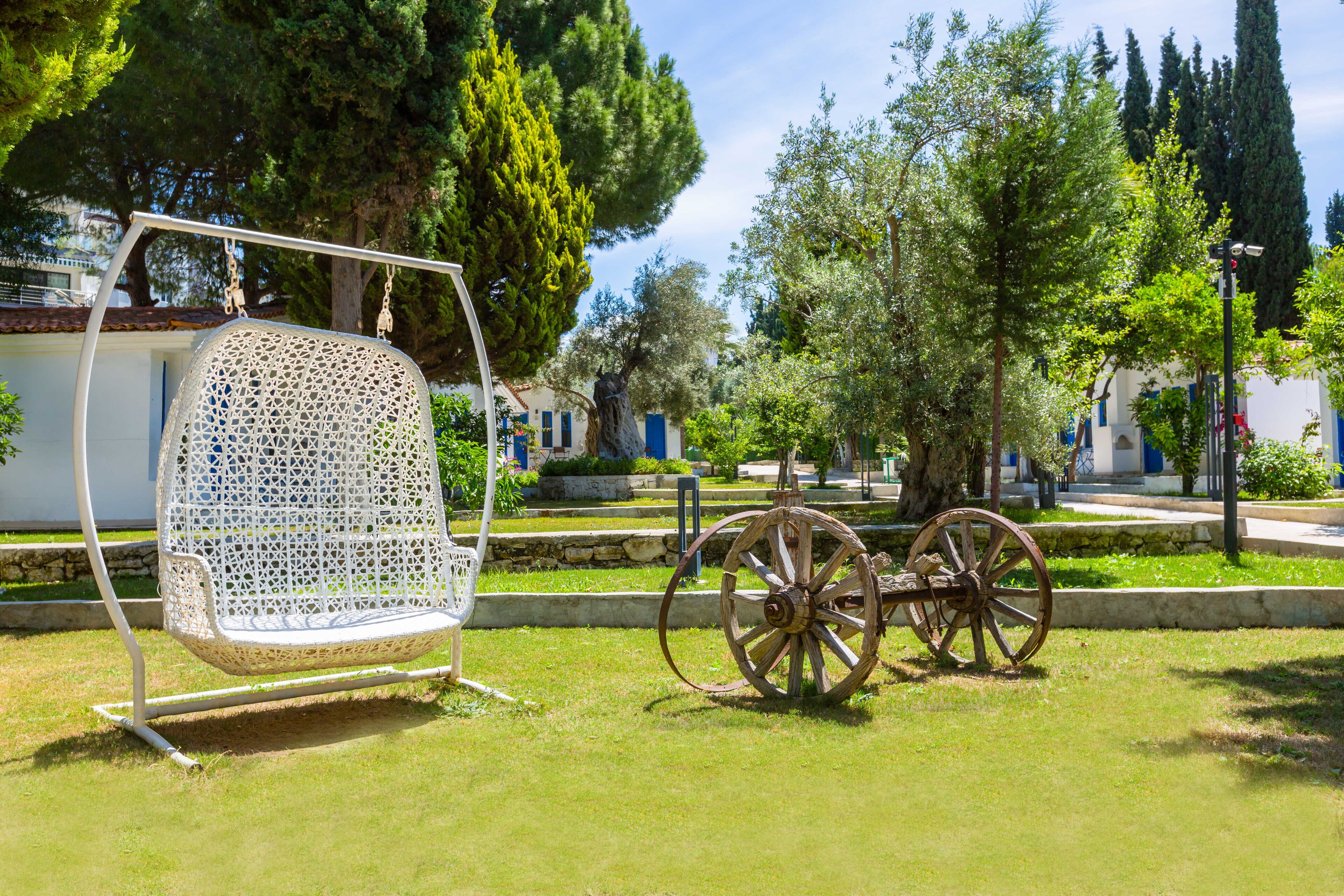 Una silla mecedora de mimbre blanca y un carrito de madera en un parque con árboles y edificios al fondo.
