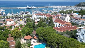 A coastal marina with numerous boats, a swimming pool surrounded by trees, and residential buildings with red-tiled roofs. - ADELLA GARDEN MARİNA HOTEL (Kusadasi)
