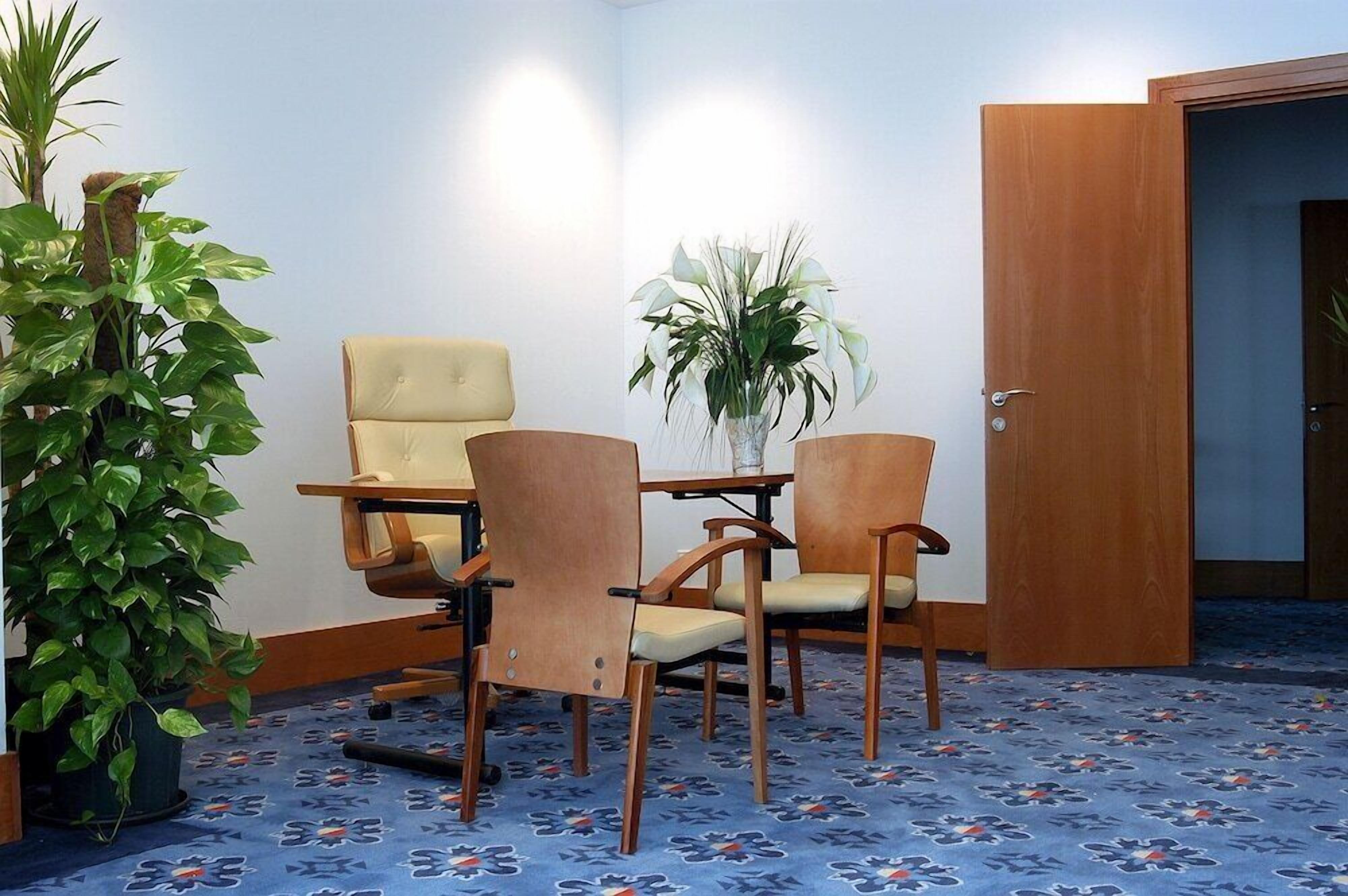 A room with a blue floral carpet, a wooden door, a beige armchair, a wooden table, and a potted plant.