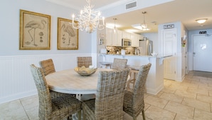 A dining area with wicker chairs, a round table, and a chandelier. There is a kitchen in the background with white cabinets and a door labeled "BEACH". - 204 Palacio, low floor professionally decorated gulf front condo!  (Pensacola)